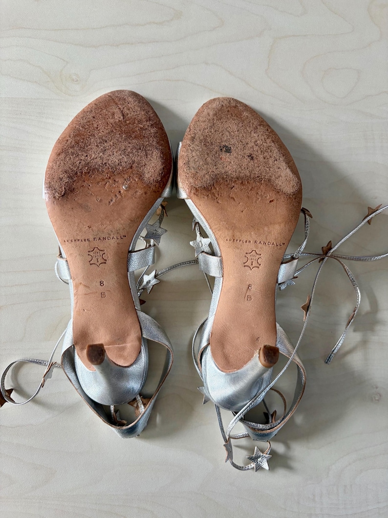 May include: A pair of silver metallic sandals with thin lace-up straps adorned with small star-shaped cutouts. The soles are tan leather with visible wear, featuring the Loeffler Randall brand name, a size 8 B stamp, and a small kitten heel.
