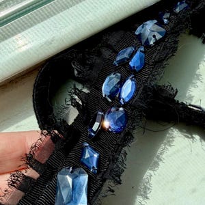 May include: Close-up of a black ribbon with frayed edges, adorned with various shapes of faceted blue gemstones. The gemstones are in different sizes and shapes, including rectangular, oval, and teardrop. The ribbon has a textured appearance.