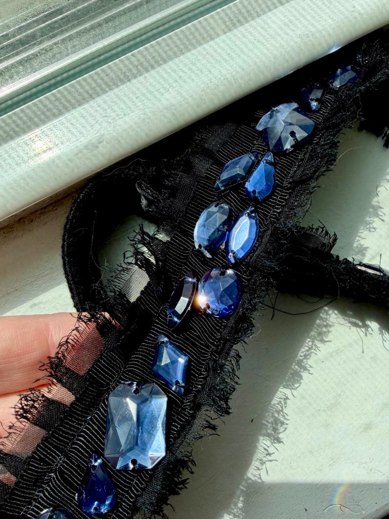 May include: Close-up of a black ribbon with frayed edges, adorned with various shapes of faceted blue gemstones. The gemstones are in different sizes and shapes, including rectangular, oval, and teardrop. The ribbon has a textured appearance.