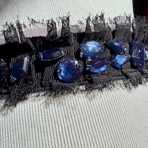 May include: A black fabric trim with a fringe edge, adorned with large, faceted blue gemstones. The trim is set against a light-colored, textured fabric, creating a contrast of textures and colors. The gemstones are various shapes and sizes.