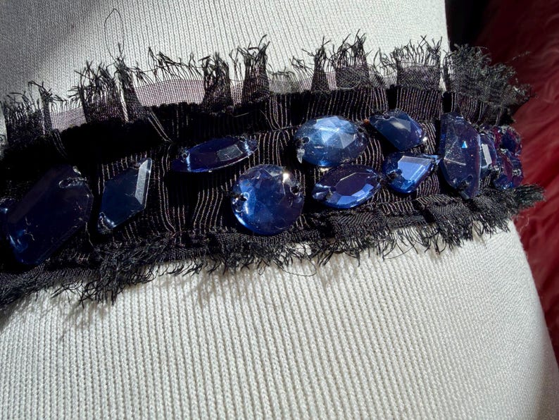 May include: A black fabric trim with a fringe edge, adorned with large, faceted blue gemstones. The trim is set against a light-colored, textured fabric, creating a contrast of textures and colors. The gemstones are various shapes and sizes.