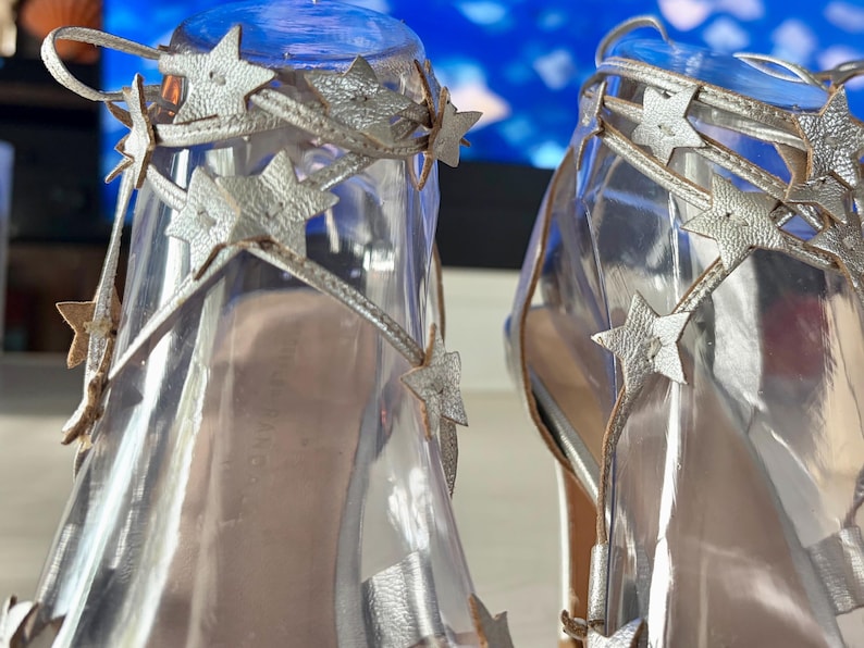 May include: A pair of silver metallic sandals featuring thin ankle straps adorned with multiple cutout star shapes. The footwear has a clear transparent material forming the upper structure, with the star-embellished straps wrapping around the ankle area.
