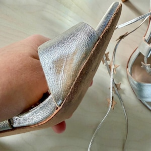 May include: A pair of metallic silver high-heeled sandals featuring a wide front strap with visible wear and discolouration on the inner edge. The sandals include long, thin silver laces adorned with small, evenly spaced metallic star-shaped charms.