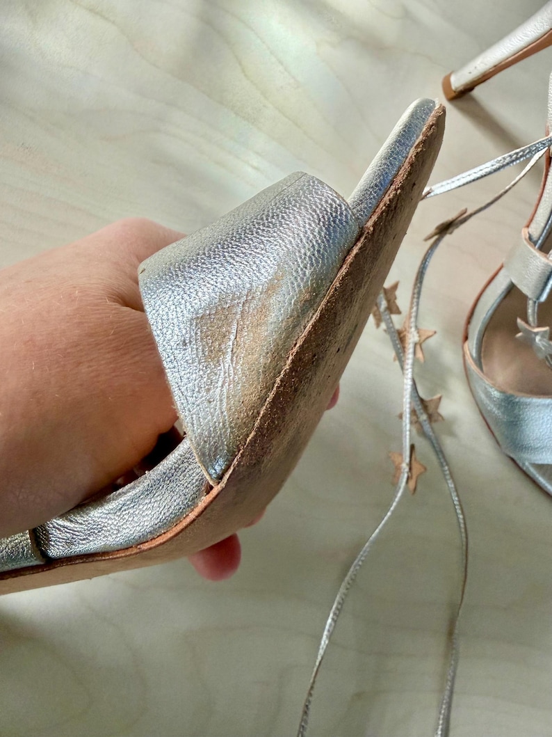 May include: A pair of metallic silver high-heeled sandals featuring a wide front strap with visible wear and discolouration on the inner edge. The sandals include long, thin silver laces adorned with small, evenly spaced metallic star-shaped charms.