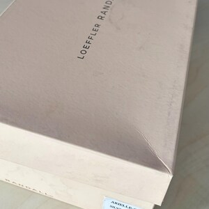 May include: A rectangular light pink cardboard shoe box with the brand name LOEFFLER RANDALL printed in black on the lid. A white rectangular label on the side displays the text ARIELLE-G, SILVER, 8B, and a barcode for product identification.