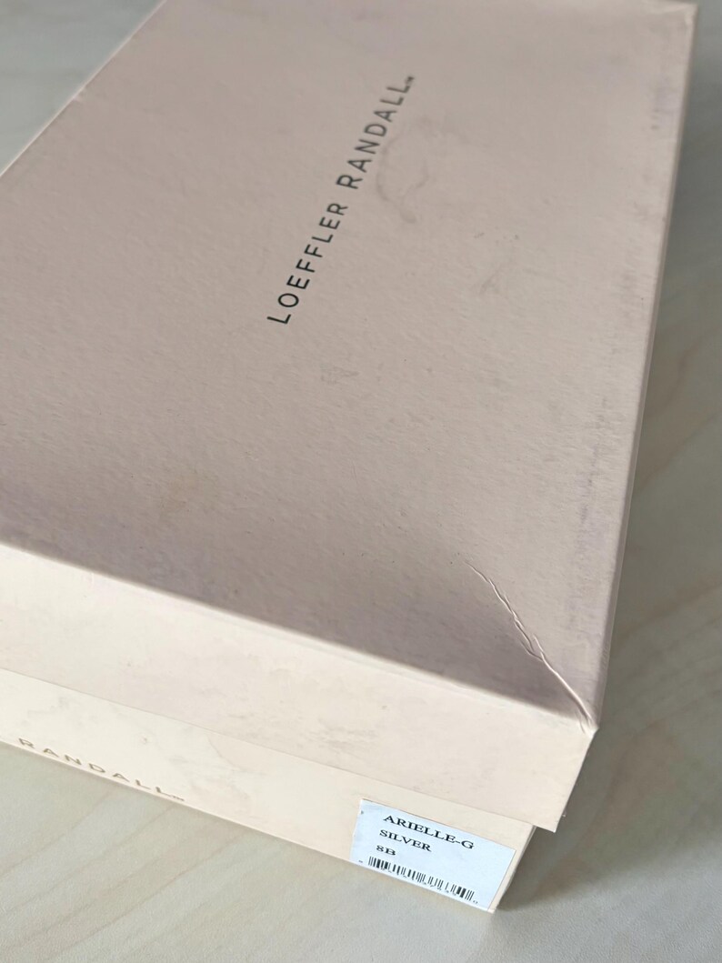 May include: A rectangular light pink cardboard shoe box with the brand name LOEFFLER RANDALL printed in black on the lid. A white rectangular label on the side displays the text ARIELLE-G, SILVER, 8B, and a barcode for product identification.