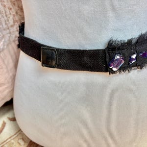 May include: Black belt with a decorative section featuring purple gemstones and a frayed edge. The belt has a black leather buckle and is worn around the waist. The belt is made of a woven material.