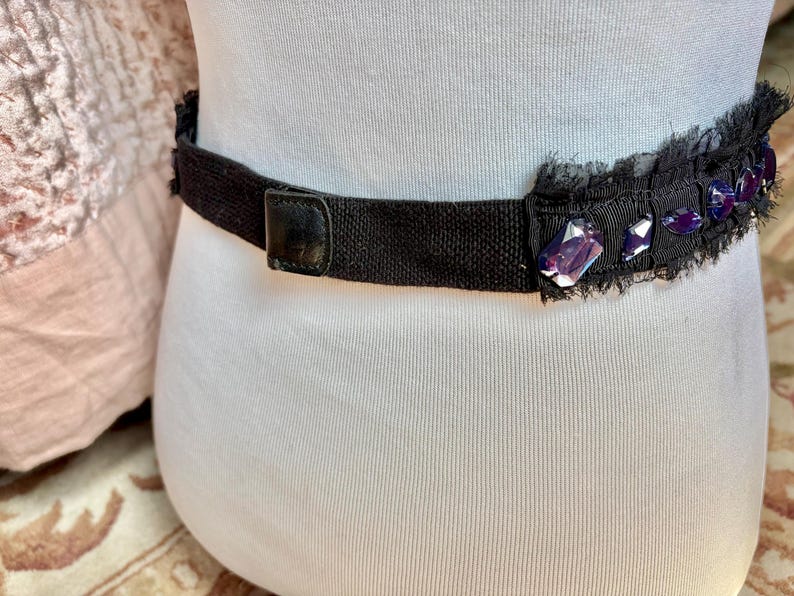 May include: Black belt with a decorative section featuring purple gemstones and a frayed edge. The belt has a black leather buckle and is worn around the waist. The belt is made of a woven material.
