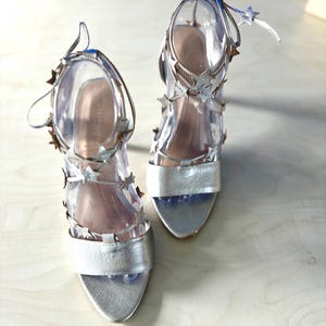 May include: A pair of silver high-heeled sandals featuring a wide toe strap and multiple thin ankle straps adorned with small metallic star-shaped embellishments. The shoes have a metallic finish and include long, slender laces that tie around the ankle.