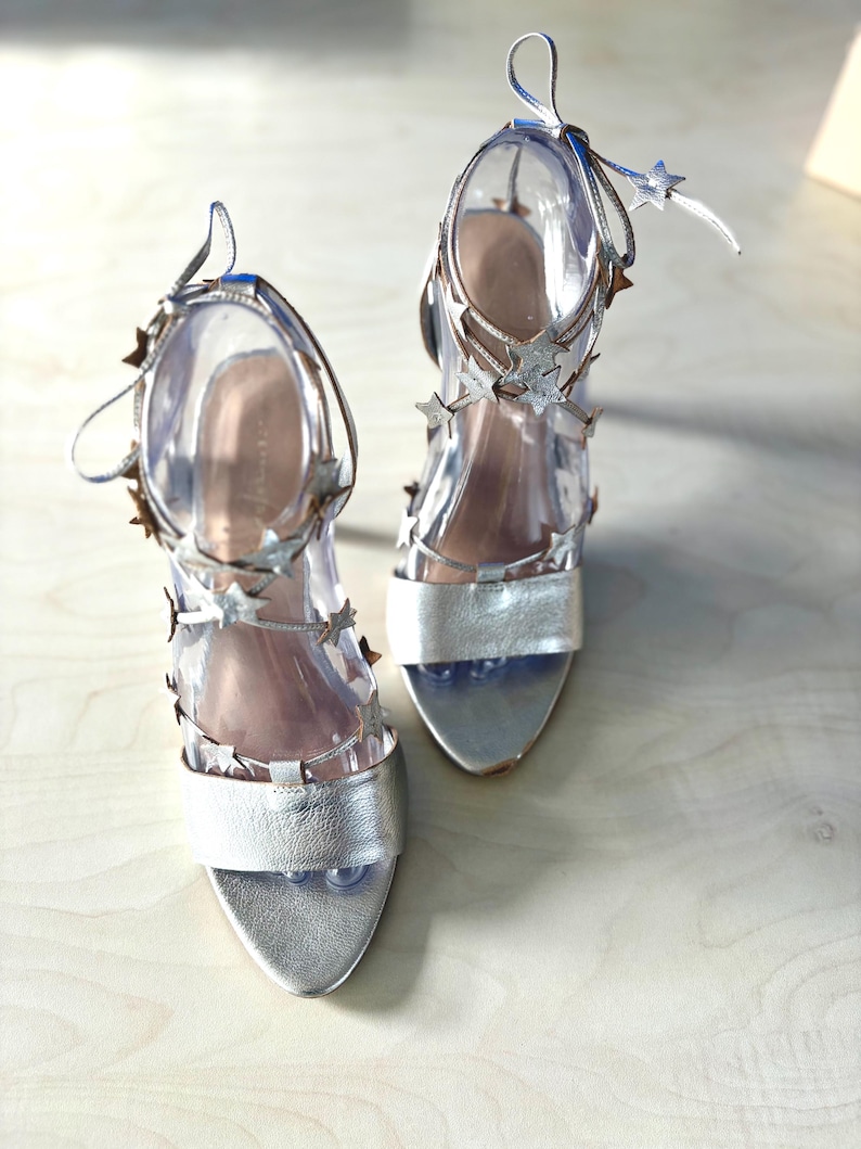 May include: A pair of silver high-heeled sandals featuring a wide toe strap and multiple thin ankle straps adorned with small metallic star-shaped embellishments. The shoes have a metallic finish and include long, slender laces that tie around the ankle.