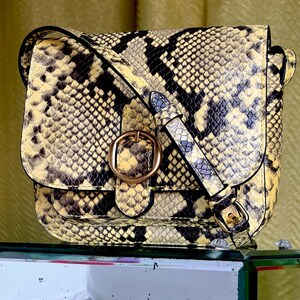 May include: A crossbody bag featuring a yellow and black snakeskin pattern. The bag has a fold-over flap secured by a circular gold-toned buckle and an adjustable shoulder strap with a matching gold-toned buckle detail on the side.