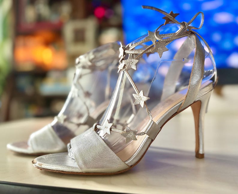 May include: A pair of silver high-heeled sandals featuring a metallic finish, an open-toe design, and thin straps adorned with multiple silver star-shaped cutouts that wrap around the ankle and tie with a bow at the back of the heel.