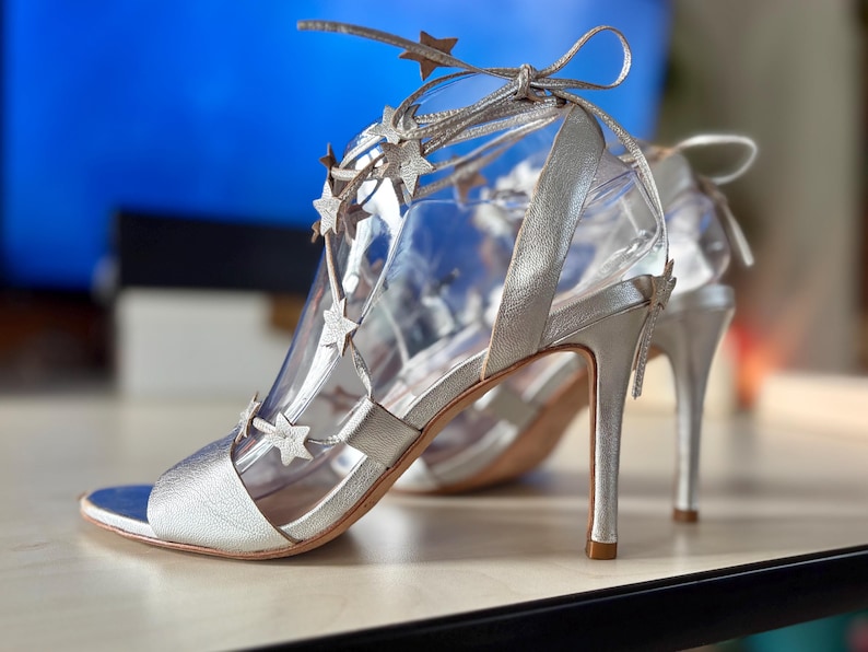 May include: A pair of metallic silver high-heeled sandals featuring a strappy design adorned with small, cut-out star embellishments. The shoes include thin laces that wrap around the ankle, a pointed toe section, and a slender stiletto heel.