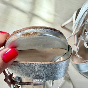 May include: A pair of metallic silver high-heeled sandals featuring lace-up straps adorned with small star-shaped cutouts. The front sole of one sandal shows visible wear and scuffing along the outer edge of the metallic silver finish.