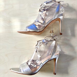May include: A pair of metallic silver high-heeled sandals featuring a peep-toe design and clear transparent side panels. The shoes are adorned with multiple small silver star-shaped embellishments along the lace-up straps that wrap around the ankle.