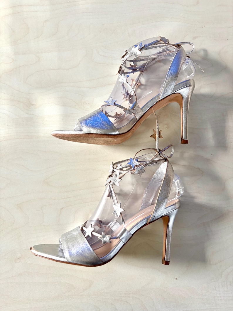 May include: A pair of metallic silver high-heeled sandals featuring a peep-toe design and clear transparent side panels. The shoes are adorned with multiple small silver star-shaped embellishments along the lace-up straps that wrap around the ankle.