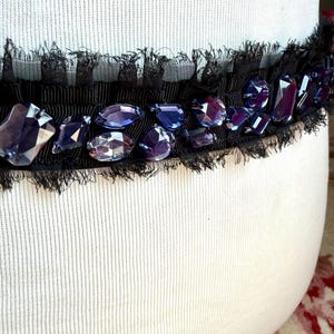 May include: Black ribbon embellished with faceted, purple-blue gemstones and a delicate fringe. The ribbon is set against a white, textured background, creating a striking contrast. This decorative trim could be used for various craft projects.