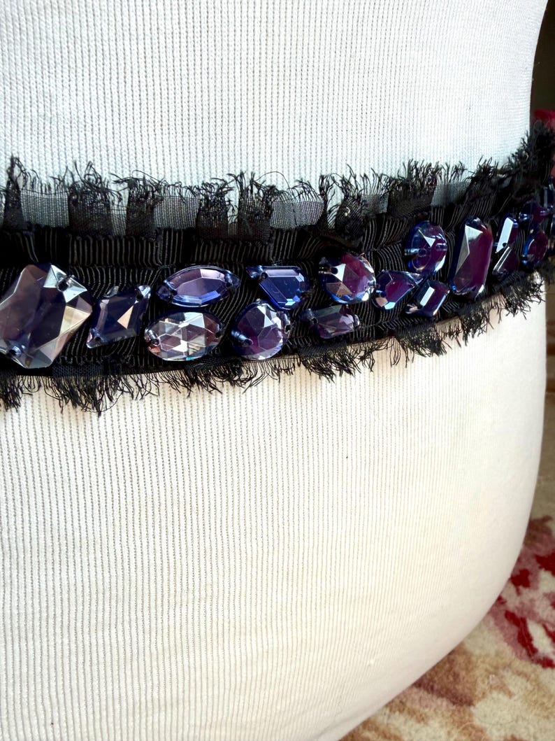 May include: Black ribbon embellished with faceted, purple-blue gemstones and a delicate fringe. The ribbon is set against a white, textured background, creating a striking contrast. This decorative trim could be used for various craft projects.
