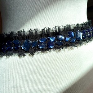 May include: A black belt with a frayed edge, adorned with irregularly shaped, dark blue gemstones. The belt is displayed on a white mannequin, showcasing its design and texture. The gemstones are of varying sizes and shapes.