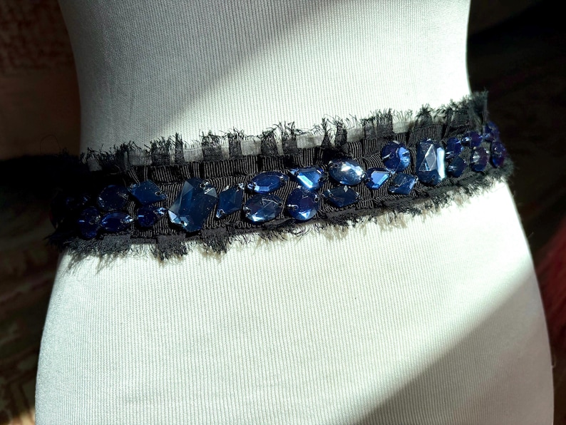 May include: A black belt with a frayed edge, adorned with irregularly shaped, dark blue gemstones. The belt is displayed on a white mannequin, showcasing its design and texture. The gemstones are of varying sizes and shapes.