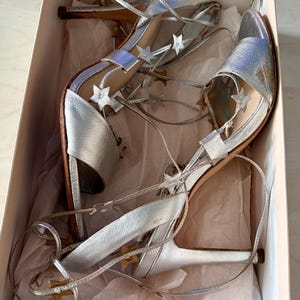 May include: A pair of silver metallic high-heeled sandals with lace-up straps adorned with small silver star cutouts. The shoes feature an open-toe design and are presented inside a light-coloured cardboard box lined with crumpled tissue paper.