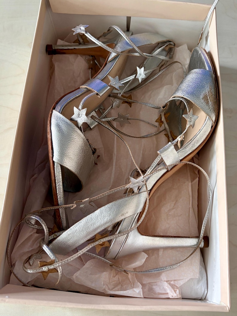 May include: A pair of silver metallic high-heeled sandals with lace-up straps adorned with small silver star cutouts. The shoes feature an open-toe design and are presented inside a light-coloured cardboard box lined with crumpled tissue paper.