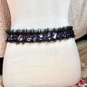May include: A black belt with a row of iridescent purple and clear gemstones. The belt has a frayed edge and is displayed on a white mannequin. The gemstones are various shapes and sizes, adding a touch of glamour.