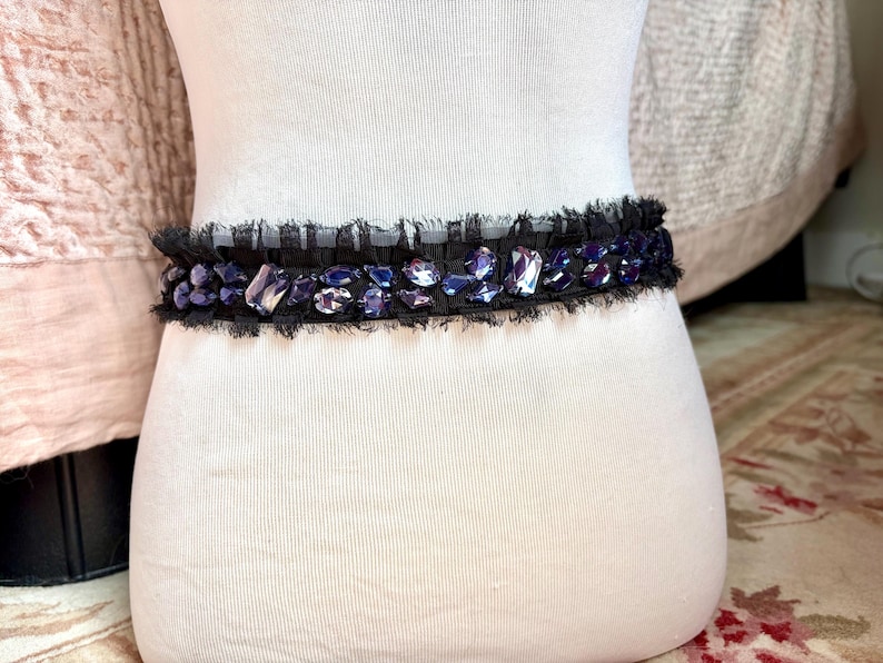 May include: A black belt with a row of iridescent purple and clear gemstones. The belt has a frayed edge and is displayed on a white mannequin. The gemstones are various shapes and sizes, adding a touch of glamour.