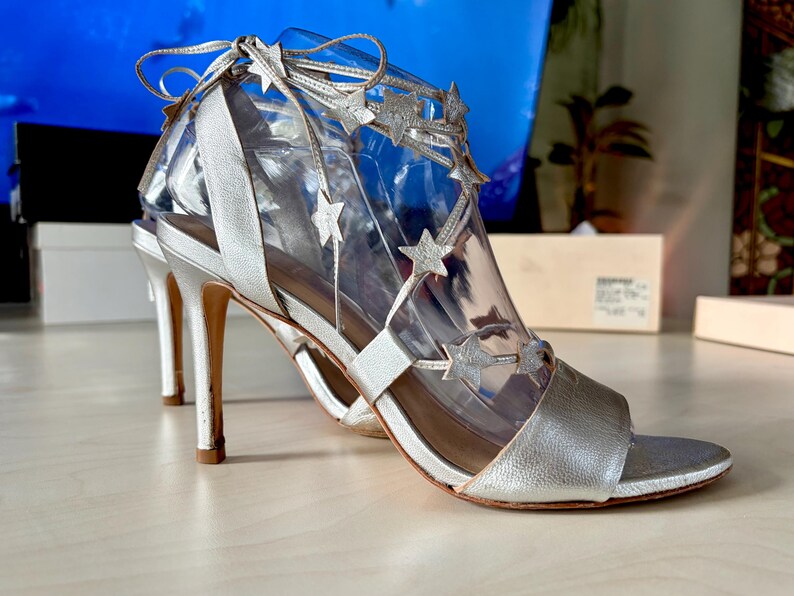 May include: A pair of metallic silver high-heeled sandals featuring a thin ankle strap that ties at the back. The upper part of the shoe is adorned with multiple silver star-shaped cutouts attached to thin straps that wrap around the foot.