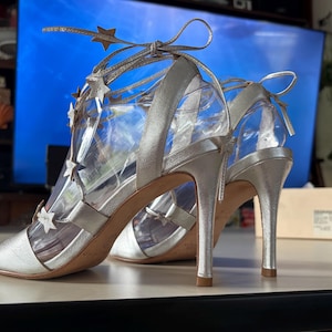 May include: A pair of metallic silver high-heeled sandals featuring a lace-up design that wraps around the ankle. The straps are adorned with multiple small silver star-shaped cutouts. The shoes have an open-toe front and a slim stiletto heel.