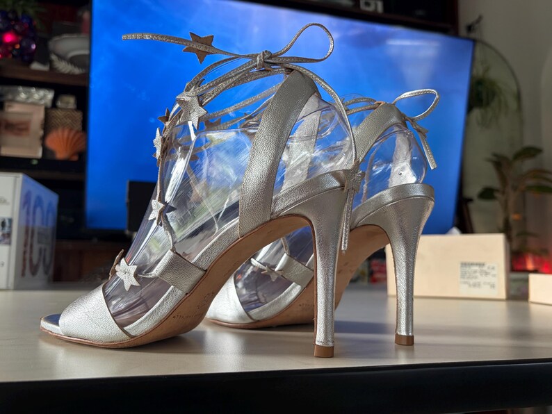 May include: A pair of metallic silver high-heeled sandals featuring a lace-up design that wraps around the ankle. The straps are adorned with multiple small silver star-shaped cutouts. The shoes have an open-toe front and a slim stiletto heel.