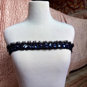 May include: A black band with blue gemstones and a ruffled edge is displayed on a white mannequin. The band is likely a decorative accessory, possibly for the neck or waist, and is set against a soft pink background.
