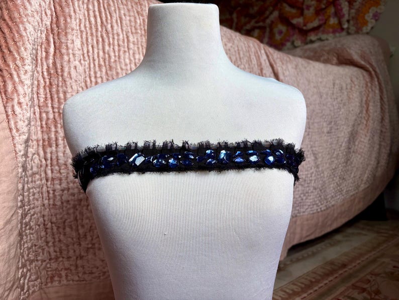 May include: A black band with blue gemstones and a ruffled edge is displayed on a white mannequin. The band is likely a decorative accessory, possibly for the neck or waist, and is set against a soft pink background.