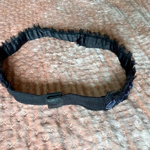 May include: A black headband with a decorative trim. The trim is made of a dark blue, fringed material. The headband has a black elastic band with a small, black leather-like tab.