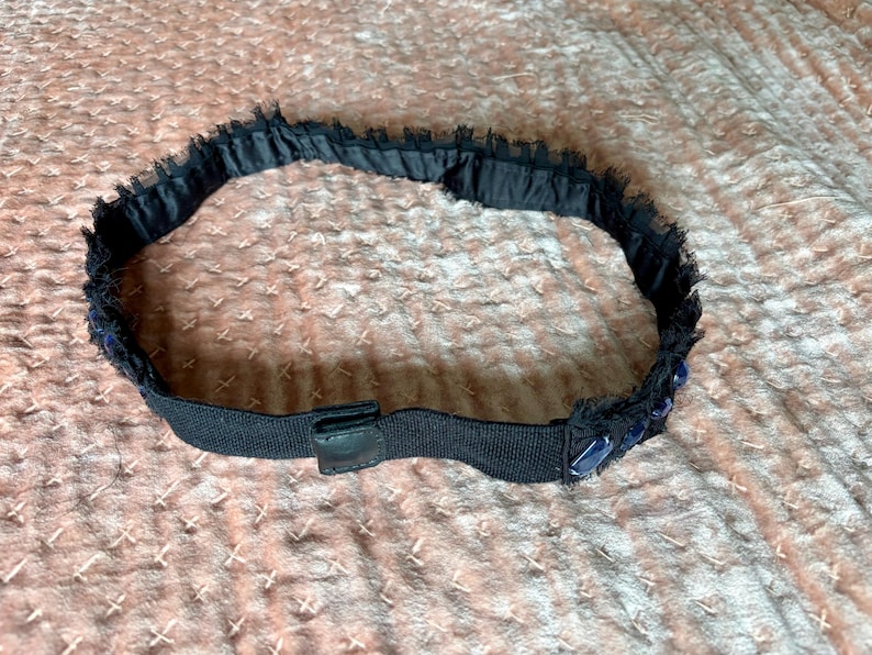 May include: A black headband with a decorative trim. The trim is made of a dark blue, fringed material. The headband has a black elastic band with a small, black leather-like tab.