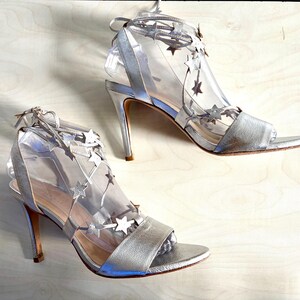 May include: A pair of silver high-heeled sandals featuring a clear transparent upper section adorned with metallic silver star-shaped cutouts. The shoes include a silver toe strap, a thin stiletto heel, and thin silver laces that tie around the ankle.