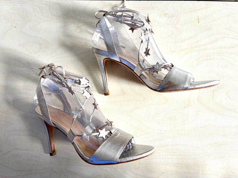 May include: A pair of silver high-heeled sandals featuring a clear transparent upper section adorned with metallic silver star-shaped cutouts. The shoes include a silver toe strap, a thin stiletto heel, and thin silver laces that tie around the ankle.