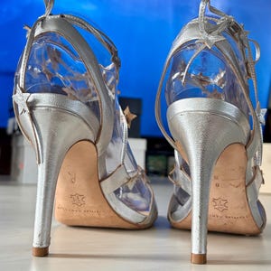 May include: A pair of silver metallic high-heeled sandals featuring thin ankle straps adorned with small star-shaped cutouts. The shoes have a stiletto heel, a tan leather sole, and a clear transparent material forming the main structure of the upper.