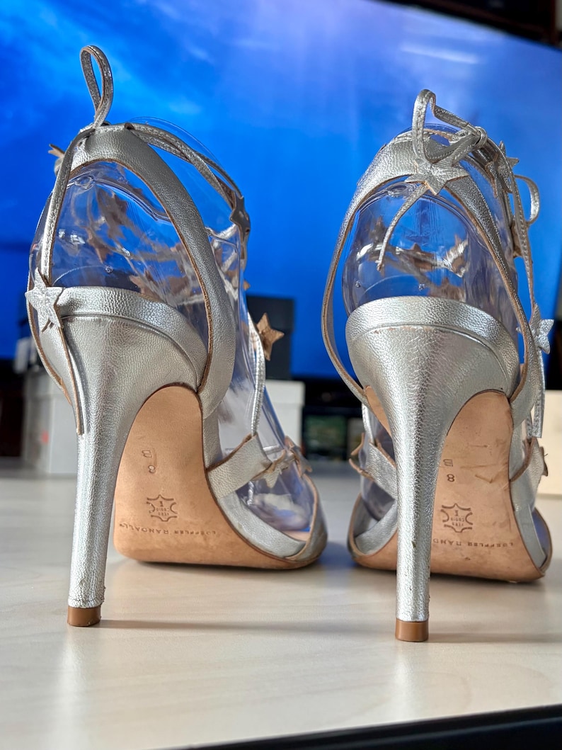 May include: A pair of silver metallic high-heeled sandals featuring thin ankle straps adorned with small star-shaped cutouts. The shoes have a stiletto heel, a tan leather sole, and a clear transparent material forming the main structure of the upper.