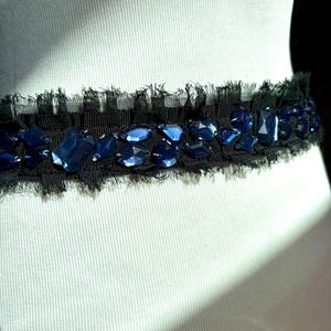 May include: A black belt with a frayed edge, adorned with irregularly shaped, deep blue gemstones. The gemstones are set against a black fabric, creating a striking contrast. The belt is displayed on a mannequin.