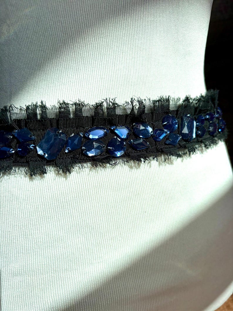 May include: A black belt with a frayed edge, adorned with irregularly shaped, deep blue gemstones. The gemstones are set against a black fabric, creating a striking contrast. The belt is displayed on a mannequin.