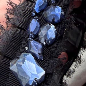 May include: Close-up of a black fabric embellished with faceted blue crystal beads. The beads are various shapes and sizes, including oval, teardrop, and rectangular. The fabric has a ribbed texture and a fringed edge.