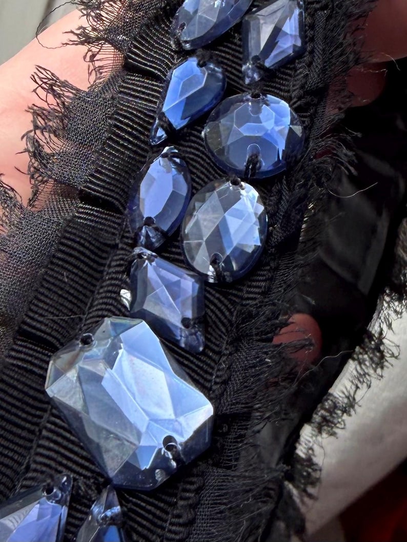 May include: Close-up of a black fabric embellished with faceted blue crystal beads. The beads are various shapes and sizes, including oval, teardrop, and rectangular. The fabric has a ribbed texture and a fringed edge.