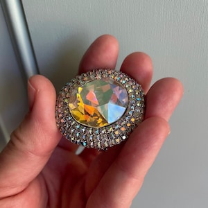 Puede incluir: Un broche grande y redondo con una piedra central facetada e iridiscente. La piedra está rodeada por un anillo de cristales más pequeños y transparentes engastados en un metal plateado. El broche se sostiene en una mano.