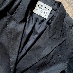 Könnte beinhalten: Ein schwarzer Blazer mit strukturiertem Finish. Das Etikett OMO Norma Kamali ist innen sichtbar. Der Blazer hat ein klassisches Revers und einreihiges Design.