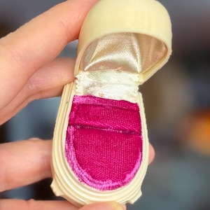 Puede incluir: Un joyero vintage abierto, de color blanquecino, con la parte superior redondeada. El interior está forrado con terciopelo rosa brillante y una tela de satén dorado. La caja está abierta por una mano.