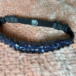 May include: Black belt with blue jewel embellishments. The belt has a black elastic band with a buckle closure. The belt is decorated with blue jewels and a black fringe.