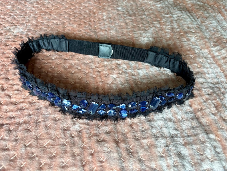 May include: Black belt with blue jewel embellishments. The belt has a black elastic band with a buckle closure. The belt is decorated with blue jewels and a black fringe.