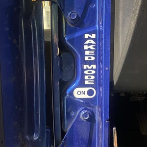 May include: Close-up of a blue vehicle door frame with the text "NAKED MODE ON" in white lettering. The door frame has visible hinges and bolts.