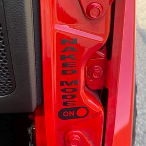 May include: Close-up of a bright red vehicle door frame. Black text reads "NAKED MODE" vertically, with an "ON" indicator below. The door frame features hinges and bolts.